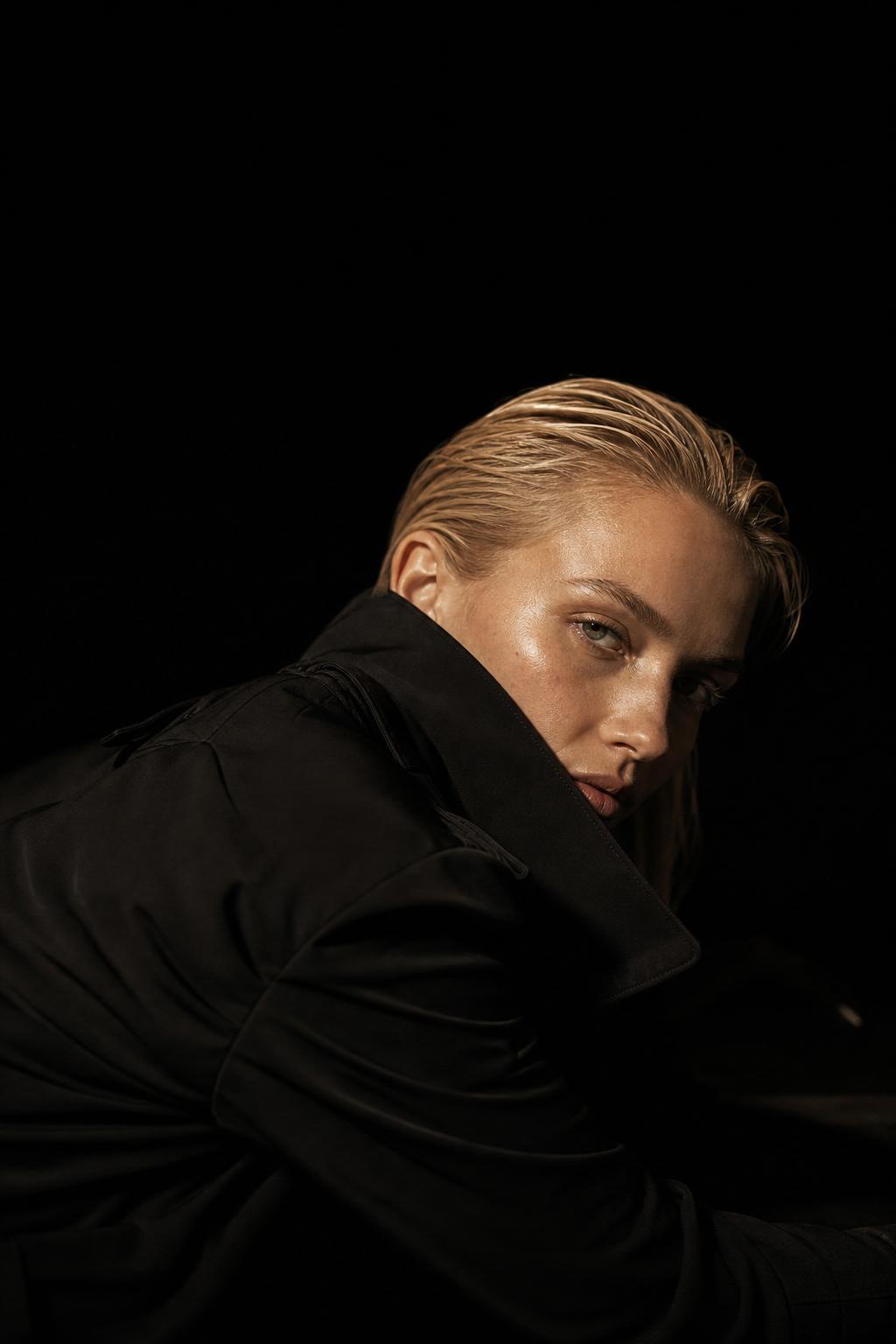 Editorial AI fashion model in a matte black trench coat reclining — Visiocy